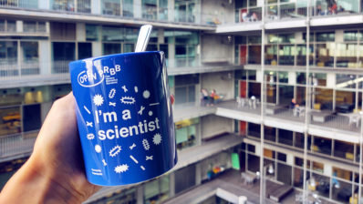 Imagen en primer plano de una taza azul con dibujos blancos donde pone I am a scientist. De fondo se ve el edificio del PRBB.