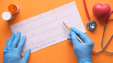 dos manos con guantes azules toman un papel con la curva del elecrtocardiograma, y ​​un bolígrafo para revisarlo. Sobre una mesa naranja también hay un fonendoscopio, un bote de pastillas abierto y un corazón pequeño de color rojo.