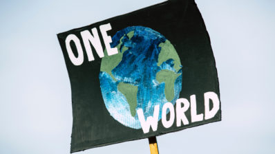 cartell pintat a mà on es veu un globus terraqui sobre fons negre i unes lletres blanques que diuen 'one world'.