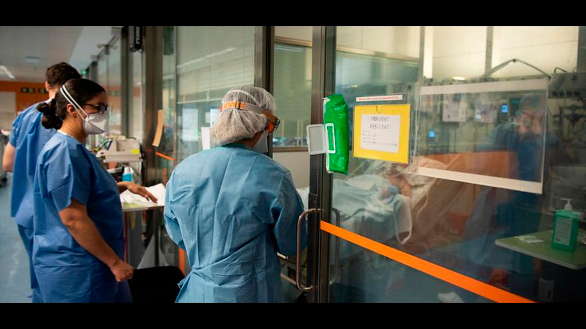 Medical staff in blue look at a patient in a glasssed-in room