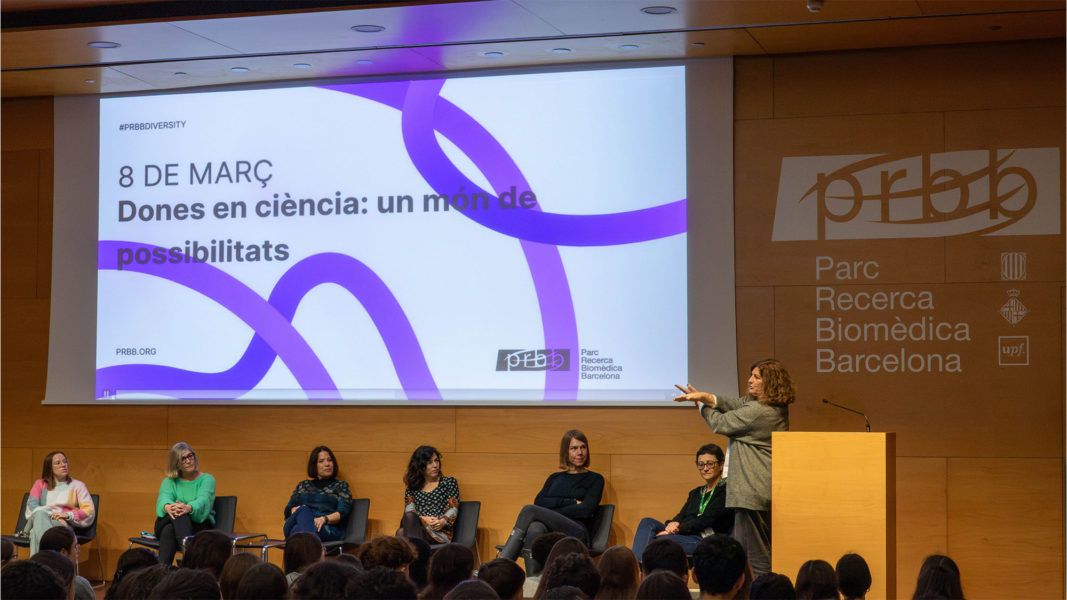 6 mujeres sentadas en el auditorio bajo una pantalla. Una mujer en el atril cruzando las manos