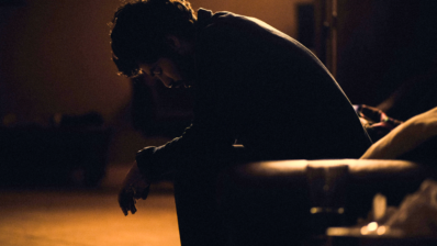 Symbolic image of a person sitting in a dark space, leaning forward with their head bowed. The warm light and strong c