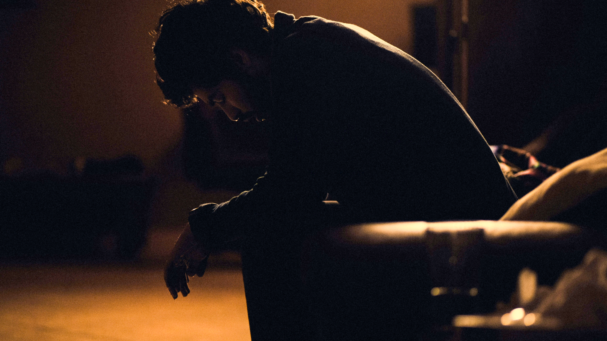 Symbolic image of a person sitting in a dark space, leaning forward with their head bowed. The warm light and strong c