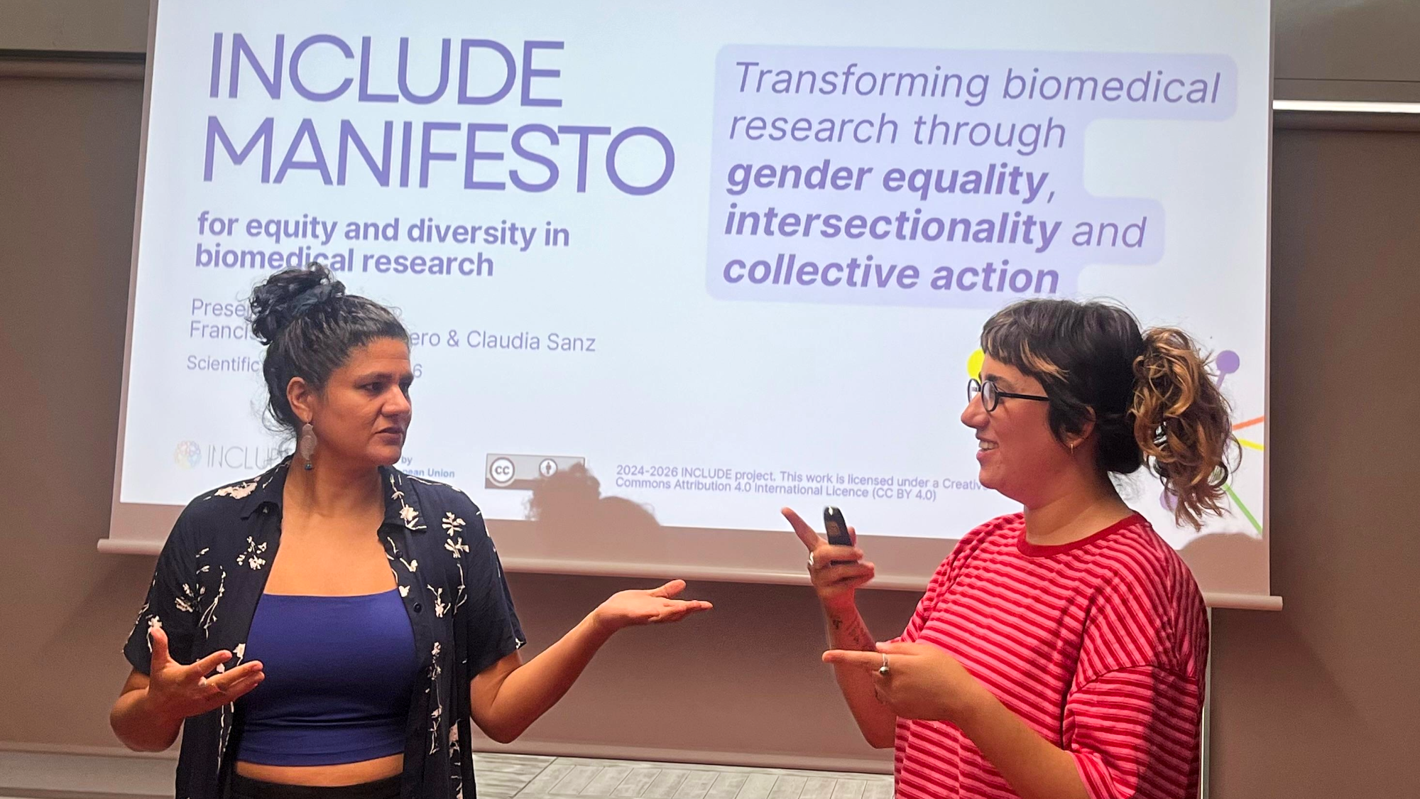 Francisca Casas-Cordero and Claudia Sanz talking in front of a screen presenting the INCLUDE Manifesto on equity and diversity in biomedical research.