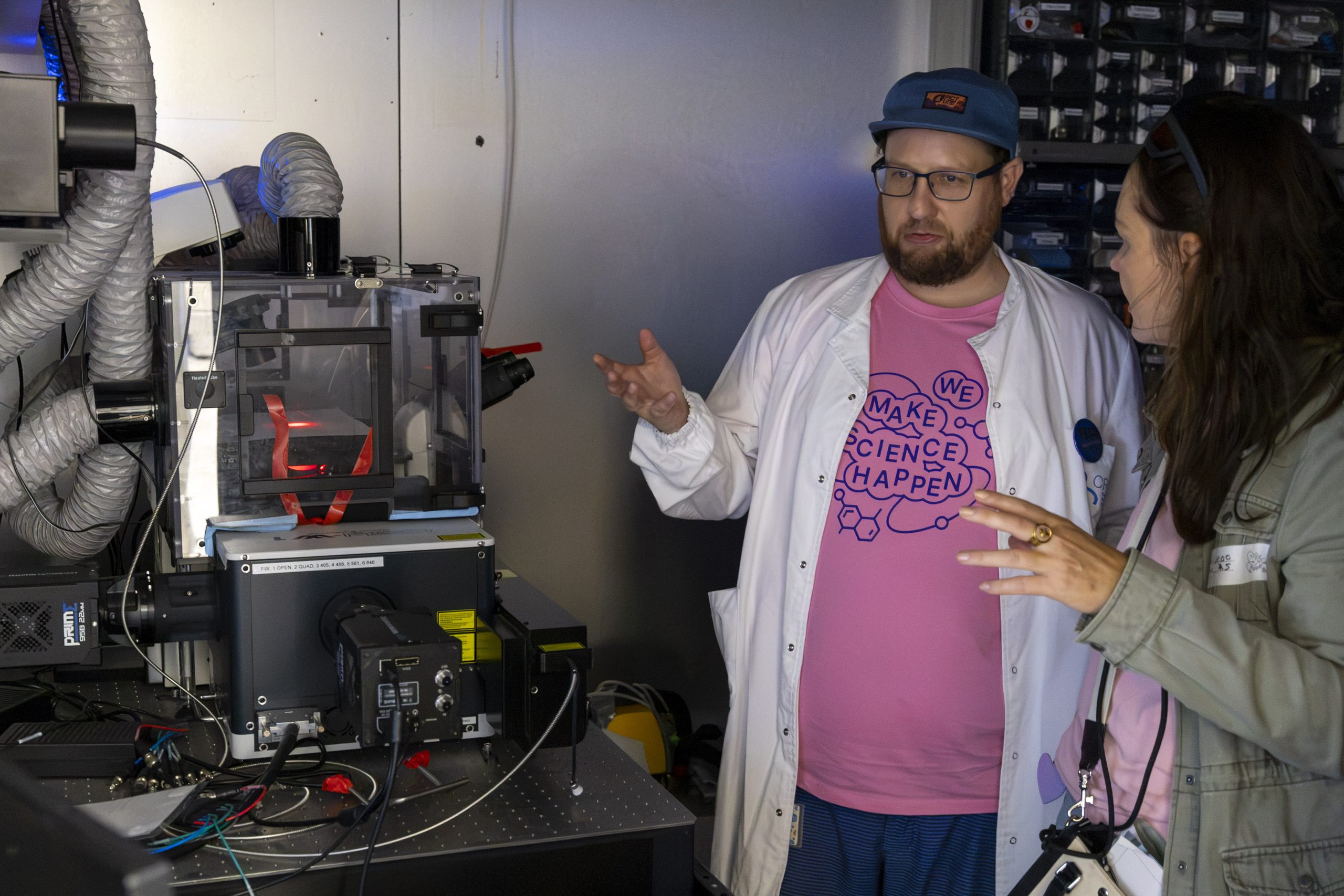 Un investigador con bata blanca y camiseta rosa de voluntario explica el funcionamiento de un equipo científico a dos visitantes durante el OpenPRBB 2026.