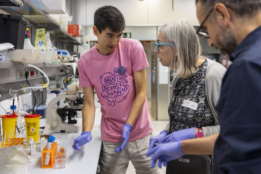 Un voluntari amb samarreta rosa i guants de laboratori mostra una activitat pràctica a dues persones visitants al costat d'un microscopi.