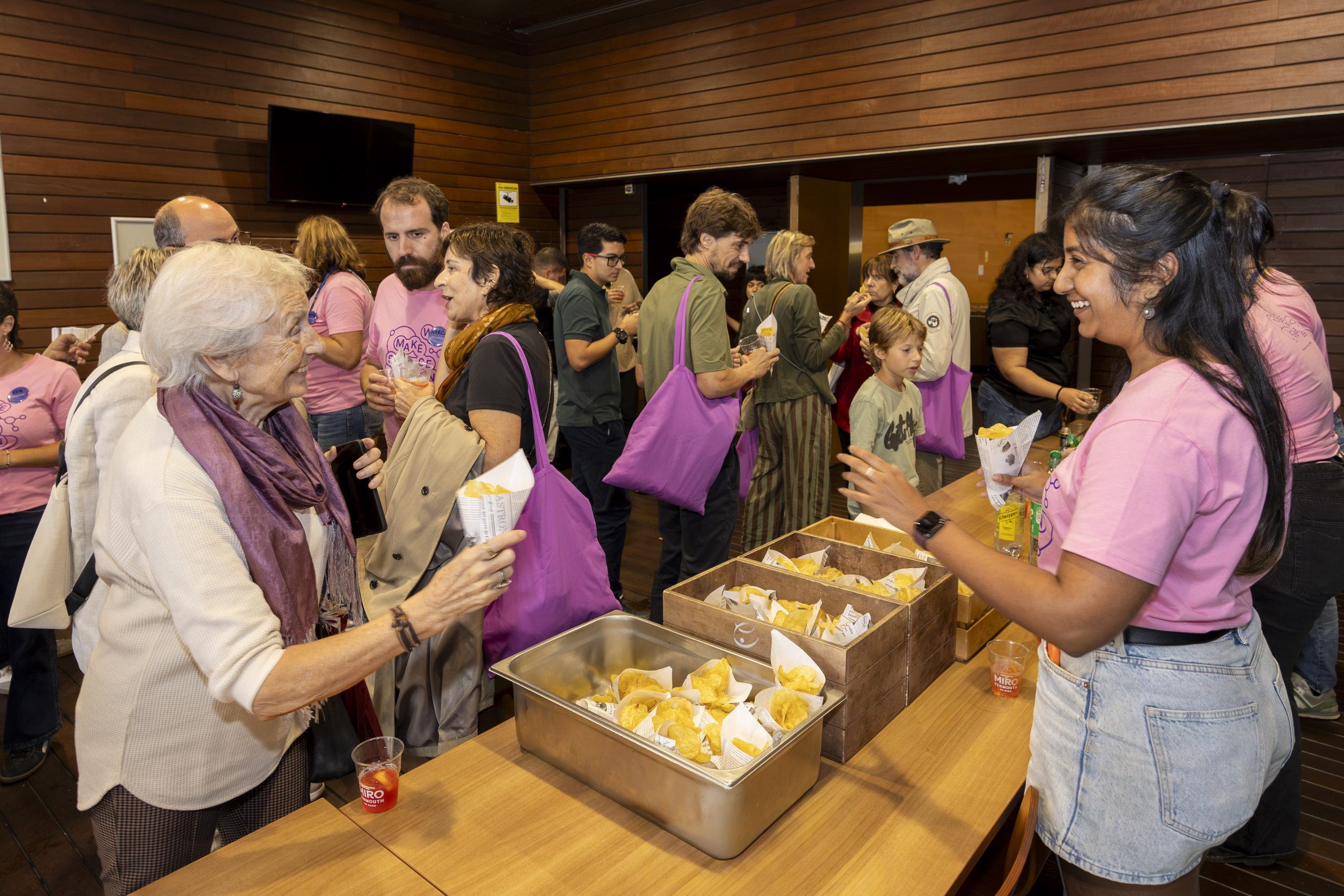 Una voluntaria con camiseta rosa, reparte aperitivos a una asistente al vermut científico del OpenPRBB 2026, mientras otros participantes conversan a su alrededor. 