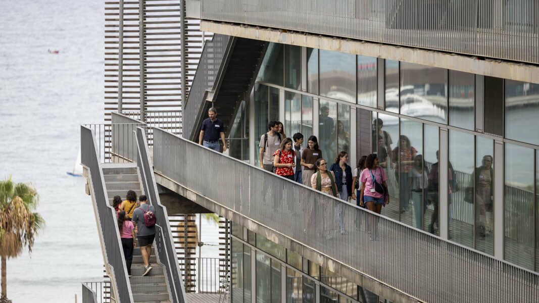 Grupo de visitantes del OpenPRBB 2026 recorriendo las pasarelas y escaleras exteriores del edificio del PRBB, con el mar de fondo.
