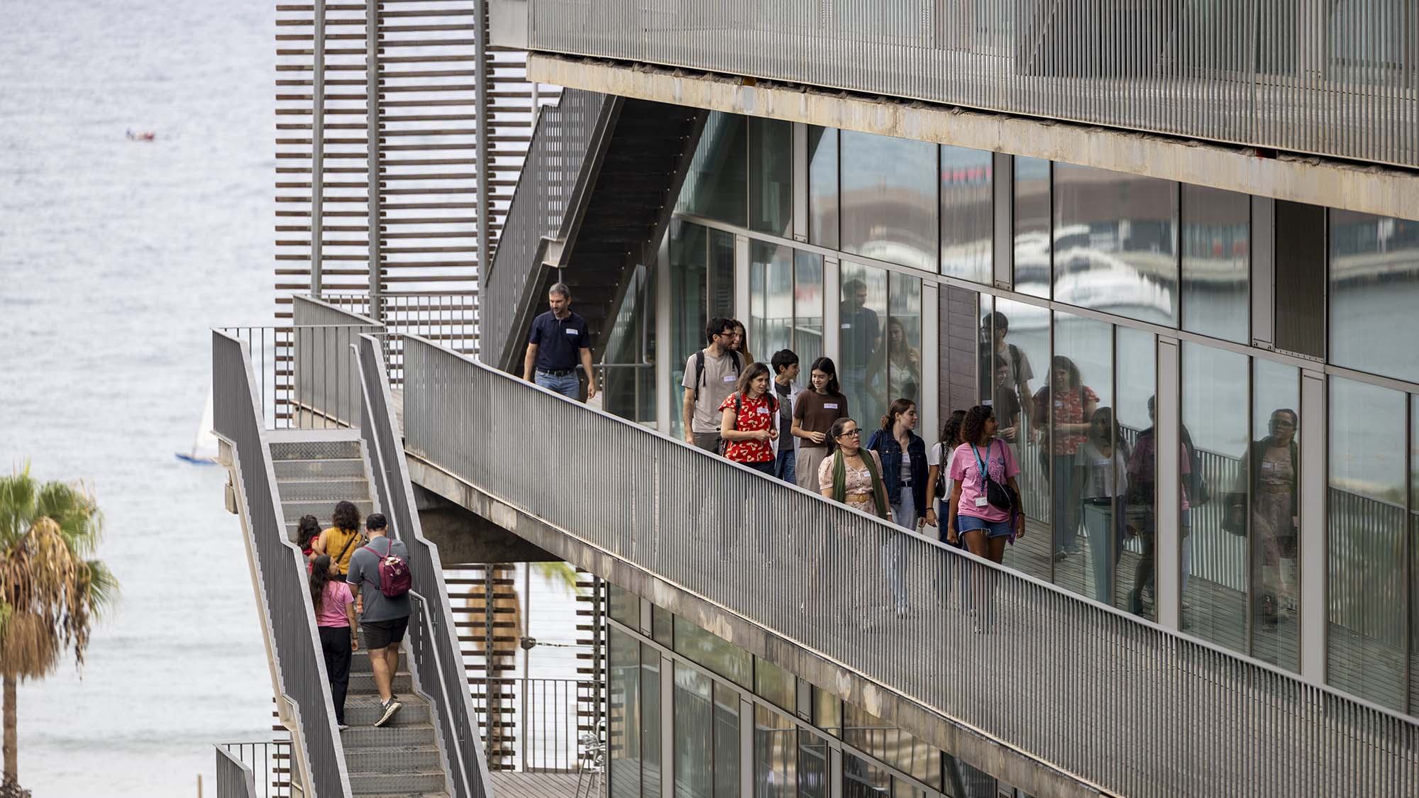 Grup de visitants de l'OpenPRBB 2026 recorrent les passarel·les i escales exteriors de l'edifici del PRBB, amb el mar de fons.