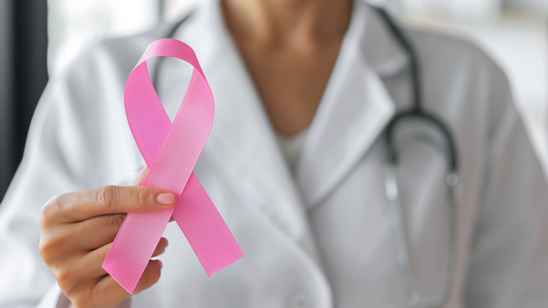A person wearing a white coat and stethoscope holds a pink breast cancer awareness ribbon in the foreground.