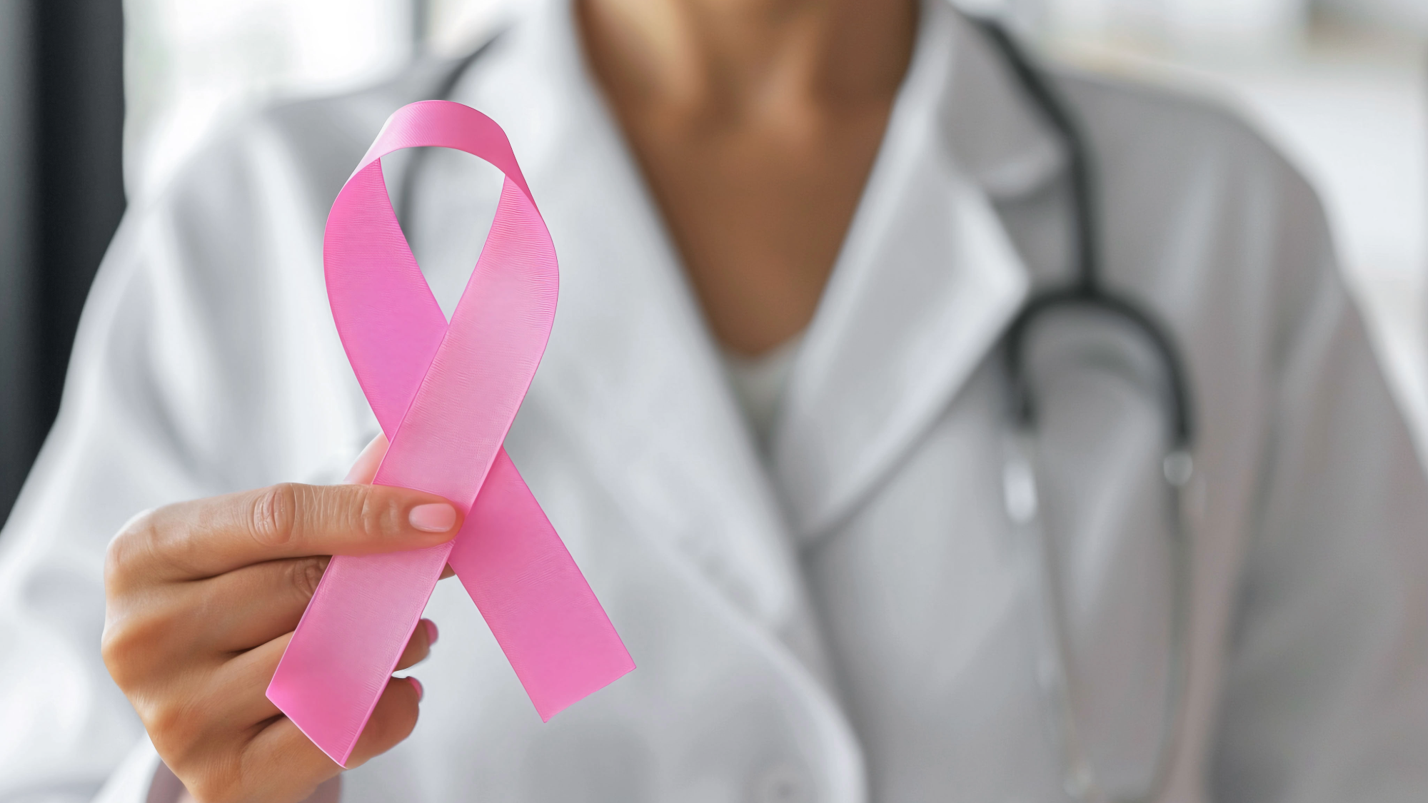Una persona con bata blanca y estetoscopio sostiene un lazo rosa, símbolo de la concienciación sobre el cáncer de mama.